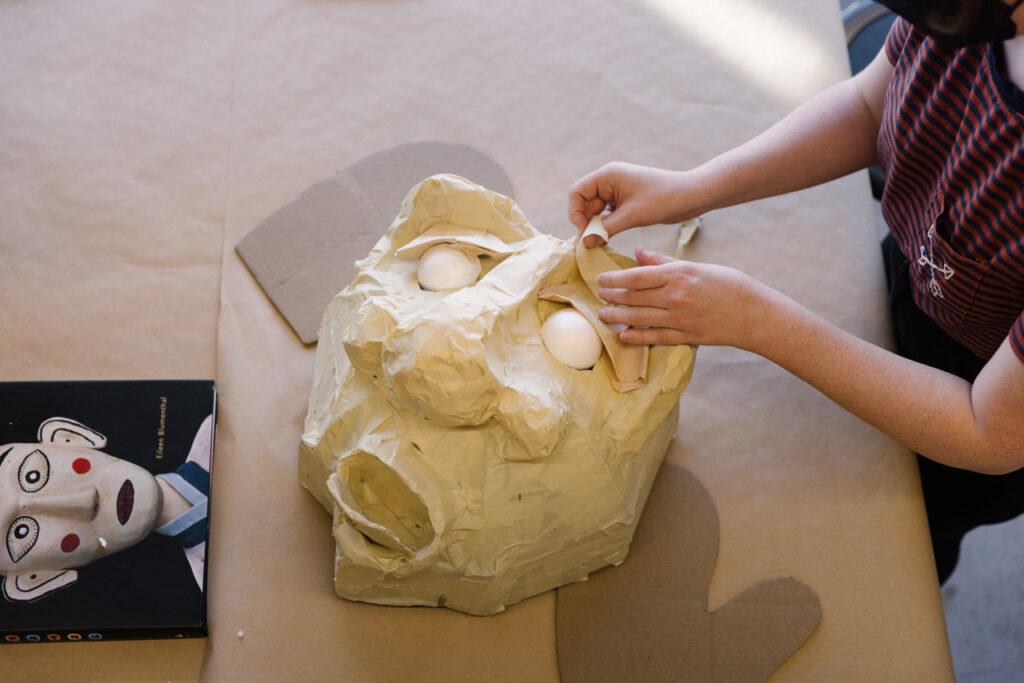 Bird's eye view of a papier mâché mask being created. From the right, a hair of hands applies another layer of paper to the mask. On the left is a reference image showing a white mask with teardrop-shaped eyes and red cheeks.