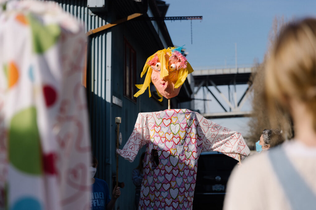 A life-sized puppet being paraded outdoors. The puppet has a blonde bob with flowers in its hair, and is wearing a pink dress with hearts. Out of focus in the foreground is another puppet's body and the back of a blonde child.