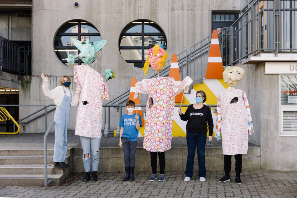 A group of people pose with large puppets outdoors. Three people are wearing puppet torsos and masks that obscure the upper half of their bodies. One puppet has a green face with large ears, the other has blonde hair and a pink face, and the third is bald with a pale face. All three are wearing dresses made of pink fabric. Beside each puppet-wearer is a person wearing a mask and leading the puppet-wearers by a stick.