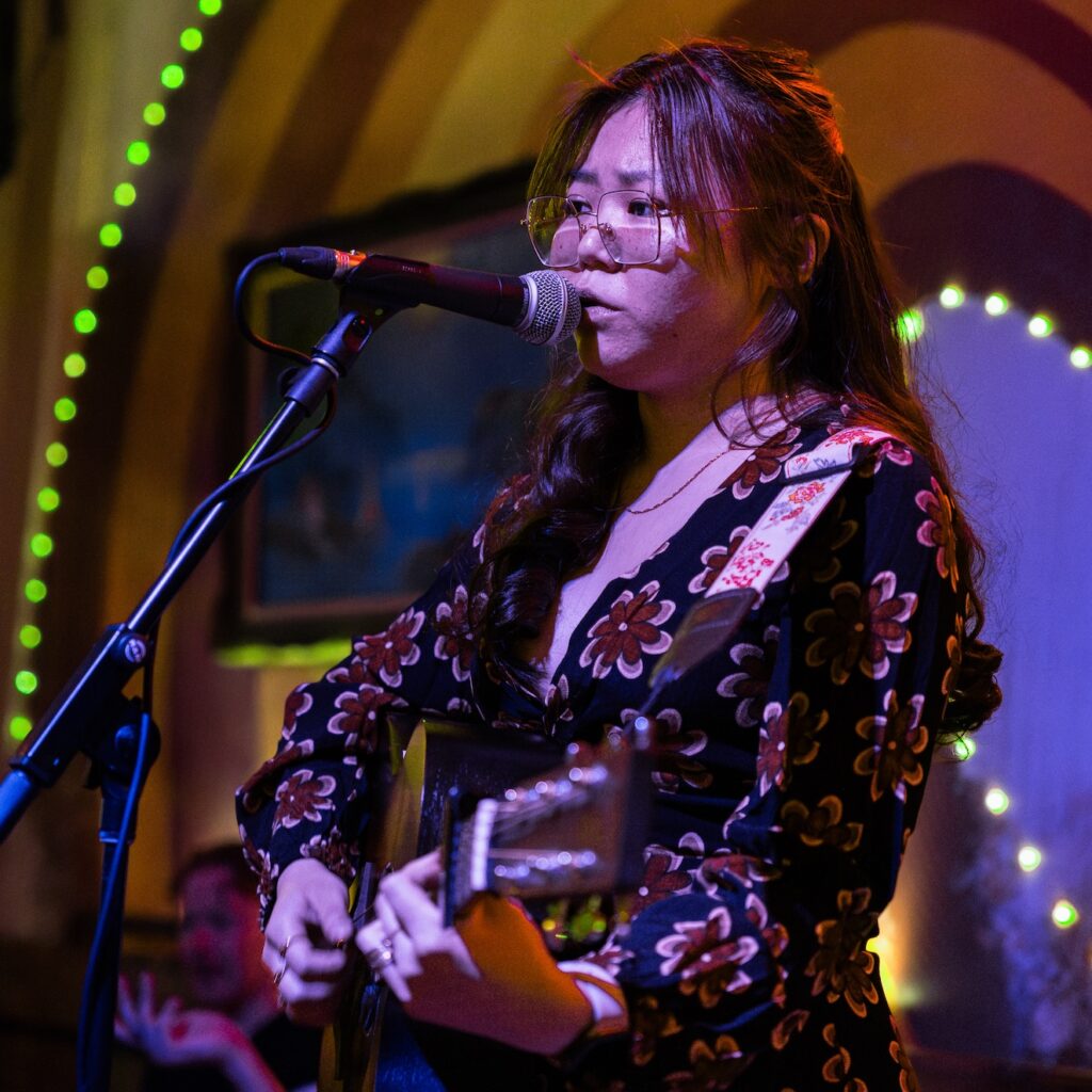 A performer wearing a floral stress, playing the guitar and singing into the microphone, bathed in pink-purple light.