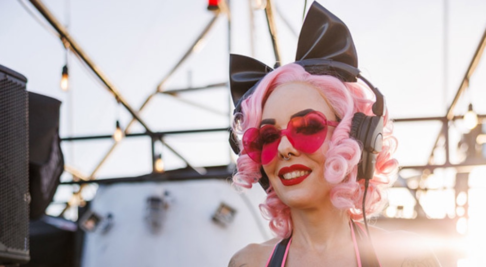 A person with curly pink hair, wearing an oversized black bow and red heart-shaped sunglasses, against an industrial outdoor background