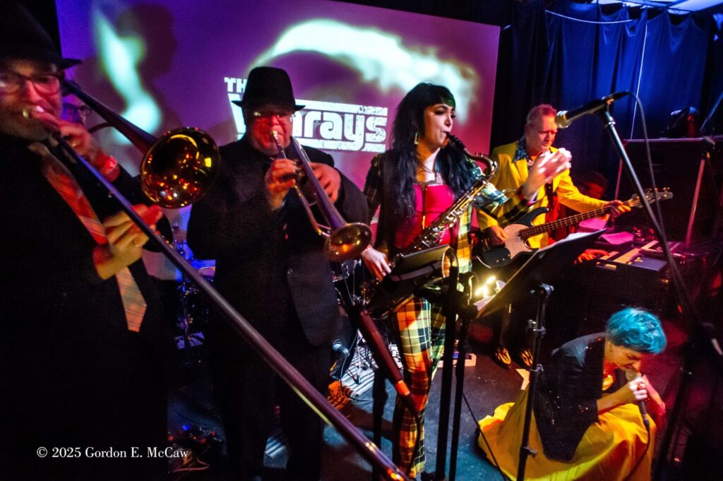 Several music performers onstage. They are playing the saxophone, trombone, and electric guitar. A vocalist squats near the ground.