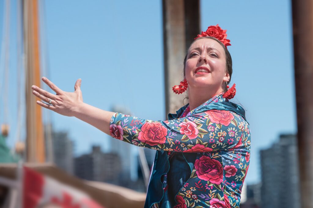 A flamenco performer with one arm oustretched. They are wearing a pink floral headband and earrings as well as a floral top. A bright blue sky fills the background.