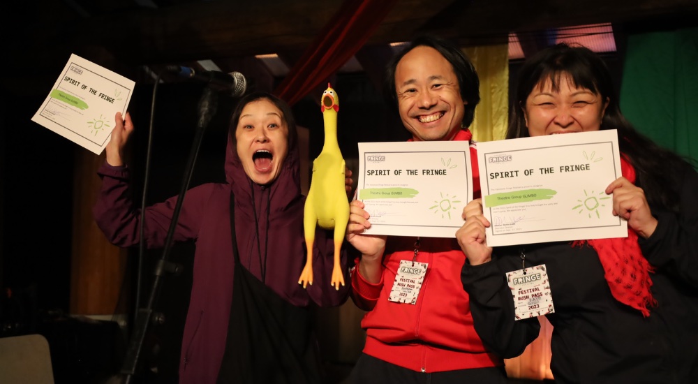 Three people holding Fringe Awards certificates to the camera. The person on the left brandishes a rubber chicken.