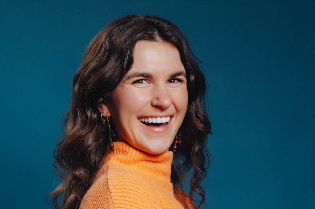 Headshot of a person with long wavy brown hair who is wearing a bright orange turtleneck. The subject is at a three-quarter angle with head turned towards the camera.