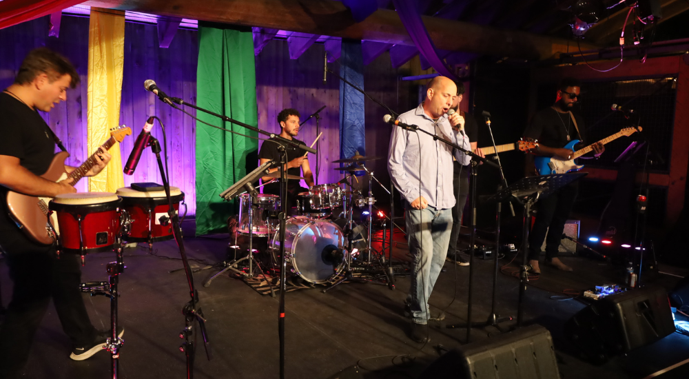 A band playing onstage with a bright purple backdrop adorned with yellow and green fabric streamers.
