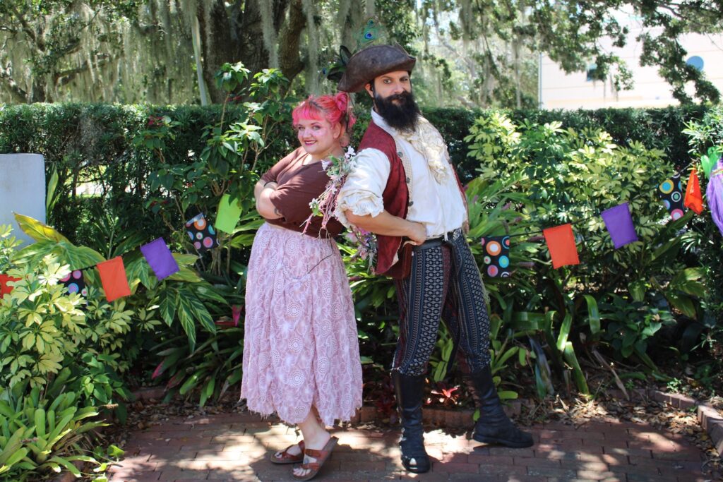 Two performers stand back-to-back, smiling at the camera. On the left, the performer has pink hair and is wearing a long pink skirt. The performer on the right is wearing a pirate costume. Behind them are hedges with coloured bunting banners strung on them.